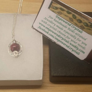 K&ouml;nnte beinhalten: Eine silberne Halskette mit einem roten Edelstein-Anh&auml;nger in einem silbernen K&auml;fig. Die Halskette wird in einer wei&szlig;en Schachtel mit einer Visitenkarte pr&auml;sentiert, auf der "Empowered Crystals" steht und Kontaktinformationen enthalten sind.