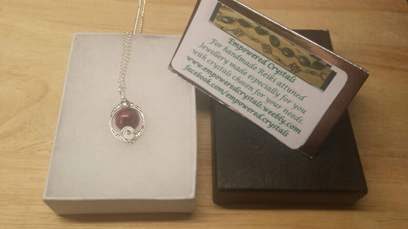 K&ouml;nnte beinhalten: Eine silberne Halskette mit einem roten Edelstein-Anh&auml;nger in einem silbernen K&auml;fig. Die Halskette wird in einer wei&szlig;en Schachtel mit einer Visitenkarte pr&auml;sentiert, auf der "Empowered Crystals" steht und Kontaktinformationen enthalten sind.