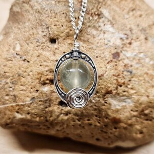 May include: A silver necklace with a pendant featuring a round, light green gemstone set in a silver frame with intricate details.