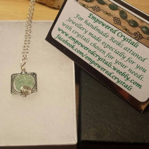 May include: A silver necklace with a green crystal pendant in a square frame. The necklace is in a white box with a black box next to it. The black box has a card with the text "Empowered Crystals" and information about the company.