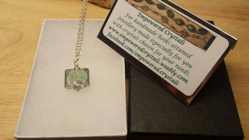 May include: A silver necklace with a green crystal pendant in a square frame. The necklace is in a white box with a black box next to it. The black box has a card with the text "Empowered Crystals" and information about the company.