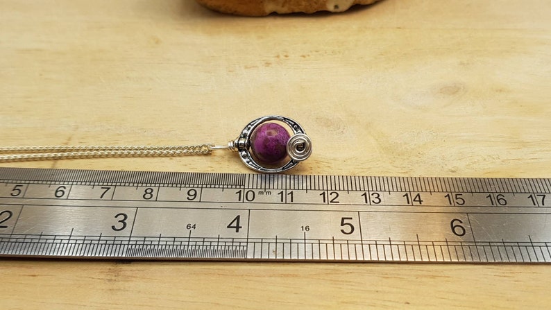 K&ouml;nnte beinhalten: Eine silberne Halskette mit einem Anh&auml;nger aus rosa und wei&szlig;em Stein. Der Anh&auml;nger ist eine Kugel mit einem silbernen Drahtdesign darum herum. Die Halskette wird an einem Lineal gemessen und ist etwa 10 cm lang.