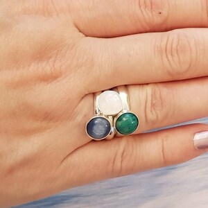 May include: A silver ring with three round gemstones: a light blue, a white, and a green stone. The ring is on a person's finger, showcasing the gemstones' colors and the ring's design. The gemstones are set in a silver band.
