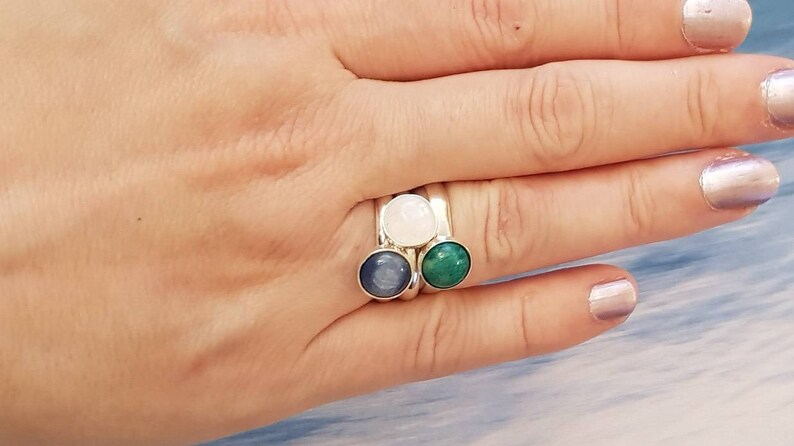 May include: A silver ring with three round gemstones: a light blue, a white, and a green stone. The ring is on a person's finger, showcasing the gemstones' colors and the ring's design. The gemstones are set in a silver band.