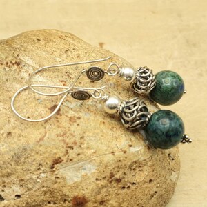May include: A pair of dangle earrings featuring dark green and blue spherical beads. The earrings have silver-colored metal accents, including decorative filigree and spiral details. The earrings are displayed on a textured, light brown stone.