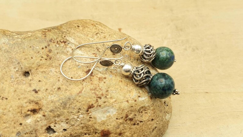 May include: A pair of dangle earrings featuring dark green and blue spherical beads. The earrings have silver-colored metal accents, including decorative filigree and spiral details. The earrings are displayed on a textured, light brown stone.