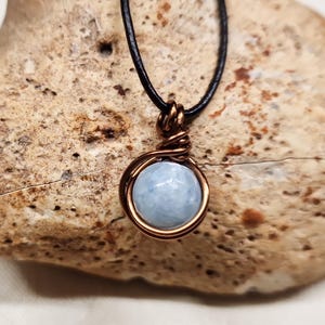May include: A round, faceted light blue gemstone pendant secured in a copper wire wrap setting. The pendant hangs from a thin black cord. The wire is coiled tightly around the top of the stone to form a loop for the cord to pass through.