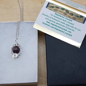 May include: A silver chain necklace with a pendant featuring a dark red gemstone encased in a silver wire design. The necklace is displayed in a white box with a black box and a card that reads "Empowered Crystals" and includes the website and Facebook page for the company.
