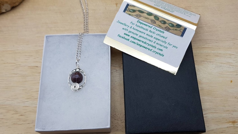 May include: A silver chain necklace with a pendant featuring a dark red gemstone encased in a silver wire design. The necklace is displayed in a white box with a black box and a card that reads "Empowered Crystals" and includes the website and Facebook page for the company.