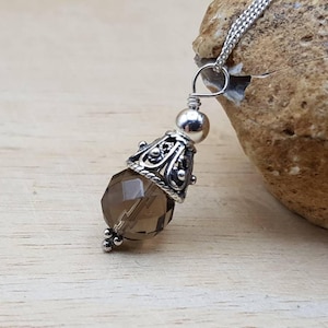 Op de afbeelding: Een zilveren hangerketting met een gefacetteerde rookkwarts edelsteen. De hanger heeft een decoratieve zilveren dop en een kleine zilveren kraal boven de edelsteen. De ketting wordt gepresenteerd op een licht houten oppervlak.