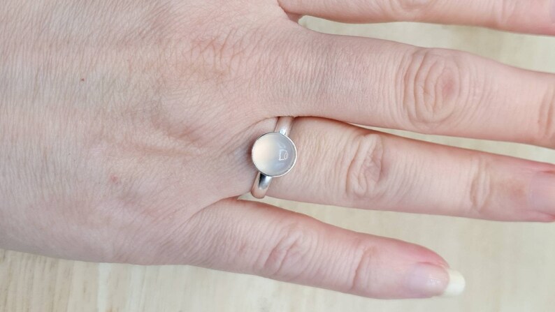 May include: A silver ring with a round, white stone set in the center. The stone has a small, engraved heart on it.