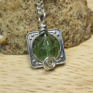May include: A silver pendant necklace with a green gemstone set in a square frame. The gemstone has a carved design.