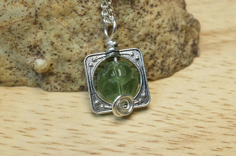 May include: A silver pendant necklace with a green gemstone set in a square frame. The gemstone has a carved design.
