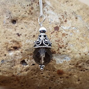 Op de afbeelding: Een zilveren hangerketting met een facetgeslepen rookkwartskristal. De hanger heeft een decoratieve zilveren dop met sierlijke filigraan en een kleine zilveren kraal aan de bovenkant. De ketting wordt getoond op een getextureerde, bruinachtige steen.