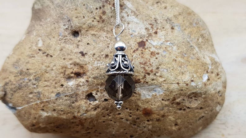 Op de afbeelding: Een zilveren hangerketting met een facetgeslepen rookkwartskristal. De hanger heeft een decoratieve zilveren dop met sierlijke filigraan en een kleine zilveren kraal aan de bovenkant. De ketting wordt getoond op een getextureerde, bruinachtige steen.