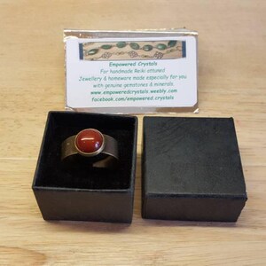 May include: A gold ring with a red gemstone set in the center. The ring is in a black box with a white label that reads "Empowered Crystals" and includes contact information.