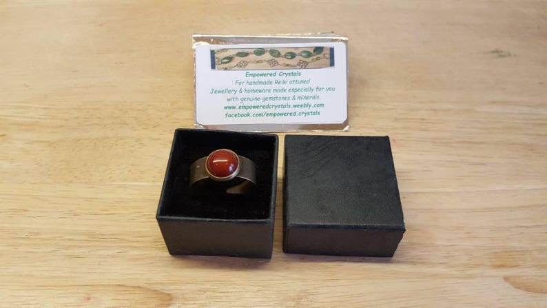 May include: A gold ring with a red gemstone set in the center. The ring is in a black box with a white label that reads "Empowered Crystals" and includes contact information.