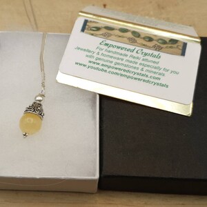 Puede incluir: Un collar con un colgante de piedra amarilla, una cadena de plata y una argolla de plata adornada. El collar est&aacute; en una caja de regalo blanca con una caja negra debajo. La caja negra tiene un borde dorado y una tarjeta que dice "Empowered Crystals" con la direcci&oacute;n del sitio web y una descripci&oacute;n de los productos.