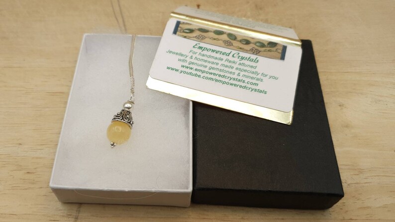 Puede incluir: Un collar con un colgante de piedra amarilla, una cadena de plata y una argolla de plata adornada. El collar est&aacute; en una caja de regalo blanca con una caja negra debajo. La caja negra tiene un borde dorado y una tarjeta que dice "Empowered Crystals" con la direcci&oacute;n del sitio web y una descripci&oacute;n de los productos.