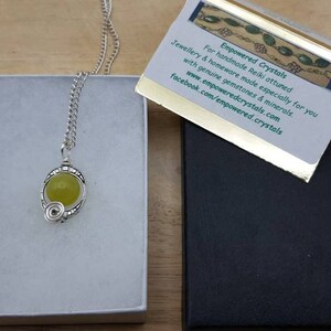 May include: A silver necklace with a green gemstone pendant in a white box. The necklace is in a silver chain and the pendant is a round green stone with a silver wire design around it. The box is on a wooden surface. The text on the card reads: "Empowered Crystals For handmade Reiki attuned jewellery & homeware made especially for you with genuine gemstones & minerals www.empoweredcrystals.com facebook.com/empowered.crystals"