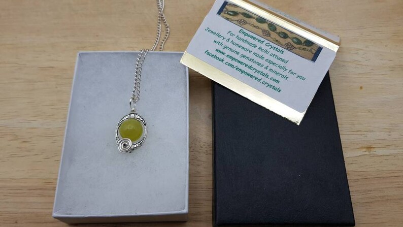 May include: A silver necklace with a green gemstone pendant in a white box. The necklace is in a silver chain and the pendant is a round green stone with a silver wire design around it. The box is on a wooden surface. The text on the card reads: "Empowered Crystals For handmade Reiki attuned jewellery & homeware made especially for you with genuine gemstones & minerals www.empoweredcrystals.com facebook.com/empowered.crystals"