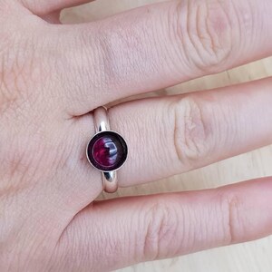 May include: A silver ring with a round, deep red gemstone set in a simple band. The gemstone is the focal point, catching the light and providing a pop of color against the silver. The ring is shown on a hand.