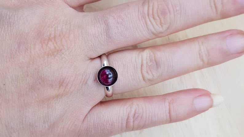May include: A silver ring with a round, deep red gemstone set in a simple band. The gemstone is the focal point, catching the light and providing a pop of color against the silver. The ring is shown on a hand.