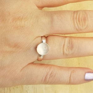 May include: A silver ring with a round, white stone set in the center. The ring is on a person's finger.