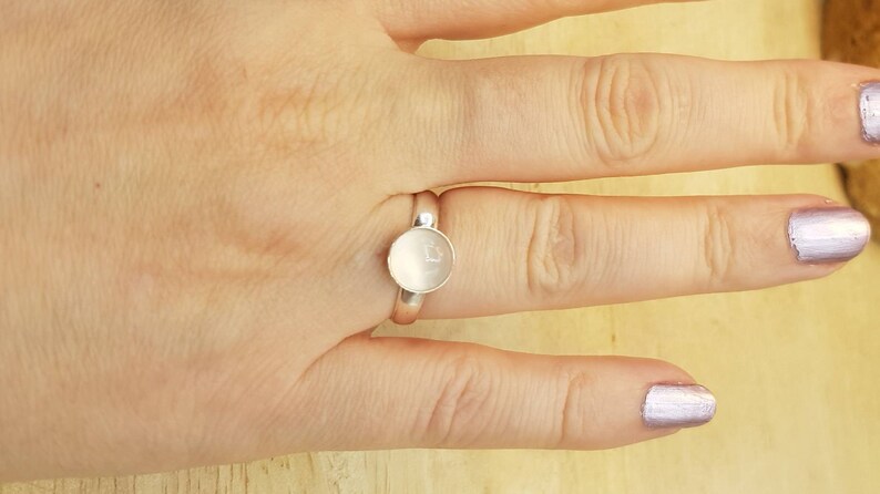 May include: A silver ring with a round, white stone set in the center. The ring is on a person's finger.