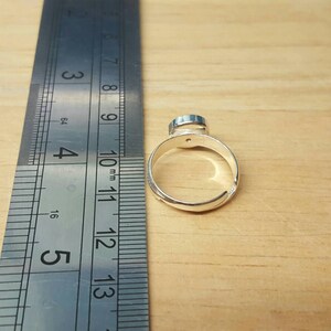 May include: A silver adjustable ring with a small, round, blue gemstone. The ring is next to a metal ruler, showing measurements in inches. The ring is set against a light brown wooden surface.
