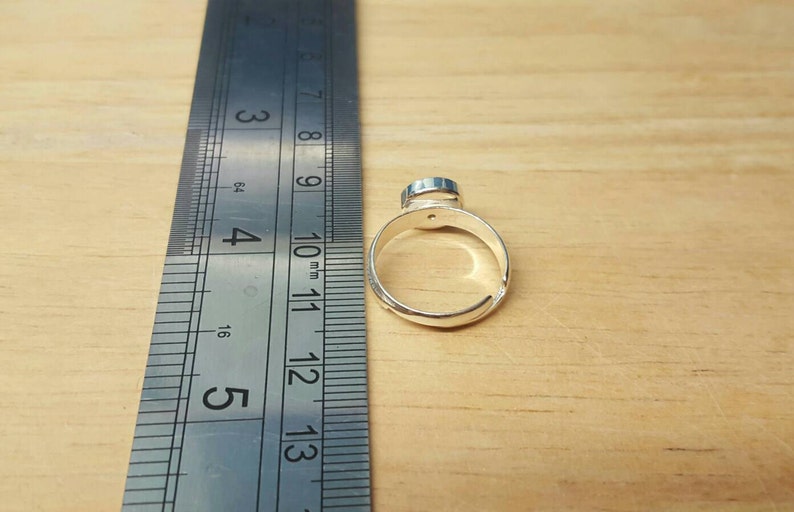 May include: A silver adjustable ring with a small, round, blue gemstone. The ring is next to a metal ruler, showing measurements in inches. The ring is set against a light brown wooden surface.
