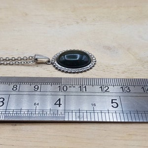 May include: A silver chain necklace with a dark green oval gemstone pendant. The pendant is surrounded by a silver beaded border. The necklace is measured against a ruler, showing the pendant is approximately 2.8 cm in length.