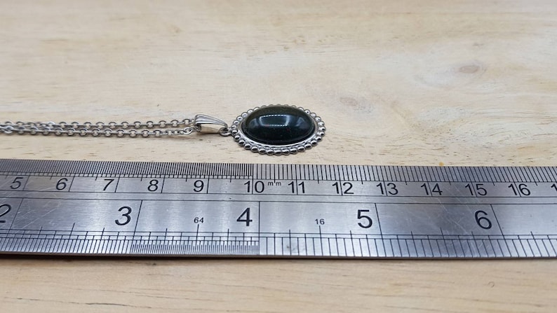May include: A silver chain necklace with a dark green oval gemstone pendant. The pendant is surrounded by a silver beaded border. The necklace is measured against a ruler, showing the pendant is approximately 2.8 cm in length.