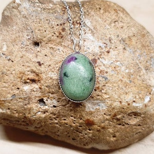 May include: A green oval pendant with a silver setting, featuring a green and pink gemstone. The pendant is hanging from a silver chain.
