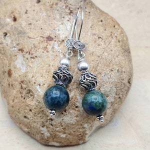 May include: A pair of dangle earrings featuring dark blue and green spherical beads. The earrings have silver-colored metal accents, including spiral details at the top and intricate cage-like designs. The earrings are displayed on a textured stone surface.