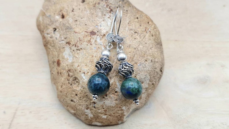 May include: A pair of dangle earrings featuring dark blue and green spherical beads. The earrings have silver-colored metal accents, including spiral details at the top and intricate cage-like designs. The earrings are displayed on a textured stone surface.