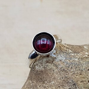 May include: A silver ring with a large, round, deep red gemstone. The gemstone is set in a bezel setting, and the ring band is smooth and polished. The ring is displayed on a textured, light brown surface.