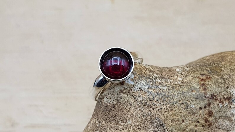 May include: A silver ring with a large, round, deep red gemstone. The gemstone is set in a bezel setting, and the ring band is smooth and polished. The ring is displayed on a textured, light brown surface.