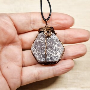 以下が含まれることがあります： 手作りのペンダントネックレス。灰と白のラフカットの石を銅線で包んでいます。ワイヤーは上部で複雑に巻かれ、ペンダントは黒いコードから吊り下げられています。石は独特の自然な質感を持っています。