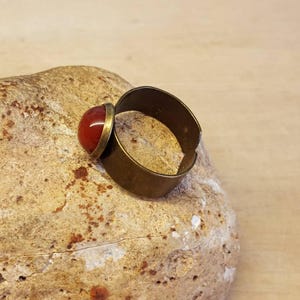 May include: A brass ring with a red gemstone set in the top. The ring is sitting on a light brown rock.
