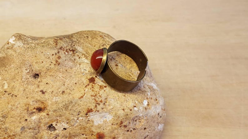 May include: A brass ring with a red gemstone set in the top. The ring is sitting on a light brown rock.