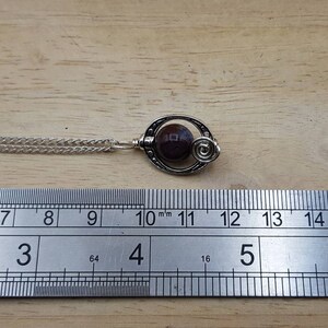 May include: A silver necklace with a pendant featuring a dark purple gemstone set in a silver swirl design. The necklace is measured against a ruler.