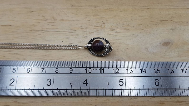 May include: A silver necklace with a pendant featuring a dark purple gemstone set in a silver swirl design. The necklace is measured against a ruler.