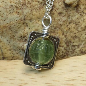 May include: A silver pendant necklace with a green gemstone set in a square frame. The gemstone has a carved design.