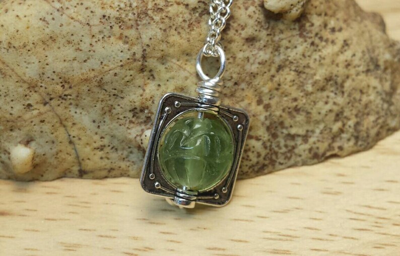 May include: A silver pendant necklace with a green gemstone set in a square frame. The gemstone has a carved design.