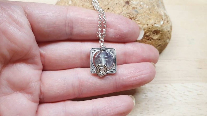 May include: A silver necklace with a square pendant featuring a clear crystal encased in wire. The pendant has a delicate silver chain.