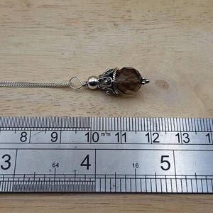 Op de afbeelding: Een zilveren hangerketting met een bruine, facetgeslepen edelsteen. De hanger heeft een diameter van ongeveer 1,3 cm en hangt aan een fijne ketting. De ketting ligt op een houten oppervlak.