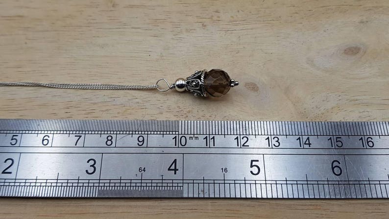 Op de afbeelding: Een zilveren hangerketting met een bruine, facetgeslepen edelsteen. De hanger heeft een diameter van ongeveer 1,3 cm en hangt aan een fijne ketting. De ketting ligt op een houten oppervlak.