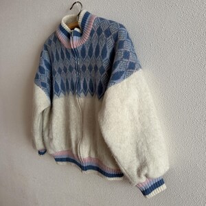 May include: A zip-up sweater with a geometric pattern in blue and white. The sweater has a high collar with pink and blue stripes, and the sleeves are a solid white. The bottom hem and cuffs have blue and pink stripes.
