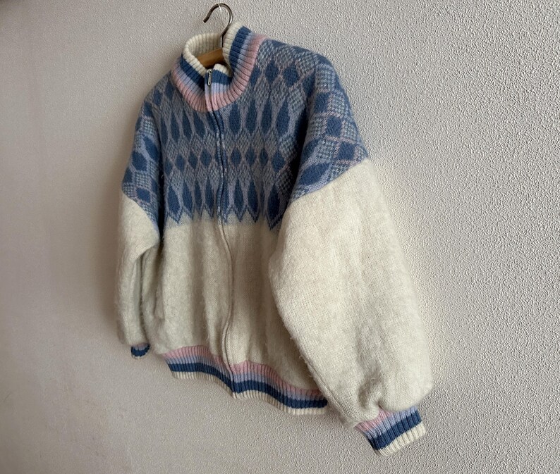May include: A zip-up sweater with a geometric pattern in blue and white. The sweater has a high collar with pink and blue stripes, and the sleeves are a solid white. The bottom hem and cuffs have blue and pink stripes.